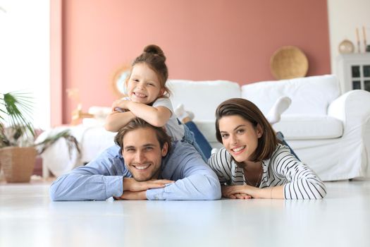 Young Caucasian family with small daughter pose relax on floor in living room, smiling little girl kid hug embrace parents, show love and gratitude, rest at home together