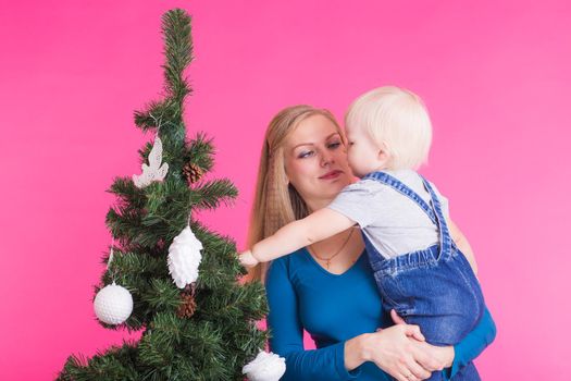 Christmas, holidays and people concept - woman and her kid near christmas tree over pink background.