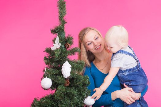 pHolidays, family and christmas concept - young woman with her baby near christmas tree on pink background.