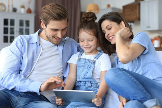Positive friendly young parents with smiling little daughter sitting on sofa together answering video call on digital tablet while relaxing at home on weekend