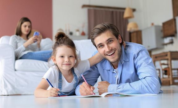 Happy father smilling daughter lying on warm floor enjoying creative activity, drawing pencils coloring pictures in albums, mother resting on couch, family spend free time together