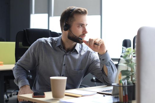 Smiling male call-center operator with headphones sitting at modern office, consulting online information in a laptop, looking up information in a file in order to be of assistance to the client