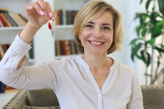 Cheerful middle aged female, real estate agent, sitting on couch, holding keys to new house