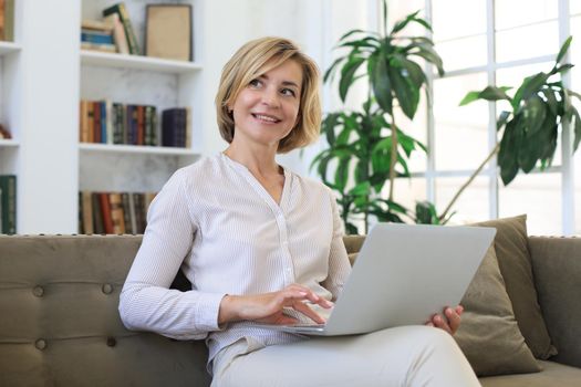 Cheerful middle aged woman using laptop while sitting on sofa at home