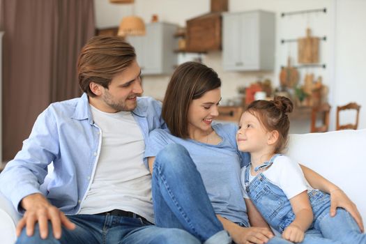 Positive friendly young parents with smiling little daughter sitting on sofa together while relaxing at home on weekend