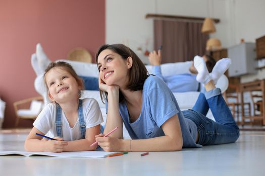 Happy mother smilling daughter lying on warm floor enjoying creative activity, drawing pencils coloring pictures in albums, father resting on couch, family spend free time together