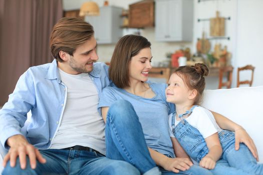 Positive friendly young parents with smiling little daughter sitting on sofa together while relaxing at home on weekend