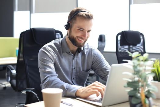 Smiling male call-center operator with headphones sitting at modern office, consulting online information in a laptop, looking up information in a file in order to be of assistance to the client
