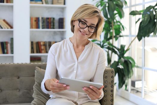 Cheerful middle aged woman sitting on sofa, using computer tablet apps, looking at screen,reading good news in social network, shopping or chatting online