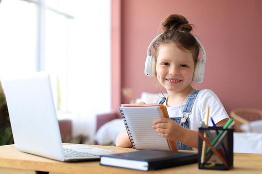 Little girl in headphones sit at desk writing in notebook studying online do exercises at home, little child handwrite prepare homework on quarantine, have web class or lesson indoors