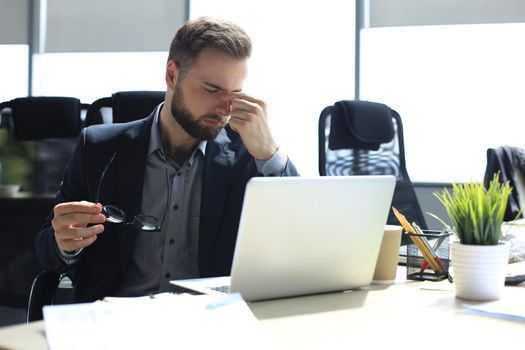Handsome young business man with closed eyes touching face with hands while sitting on working place in office