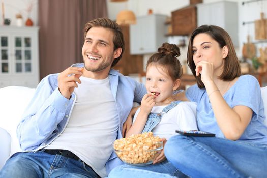 Happy family with child sitting on sofa watching tv and eating popcorn, young parents embracing daughter relaxing on couch together