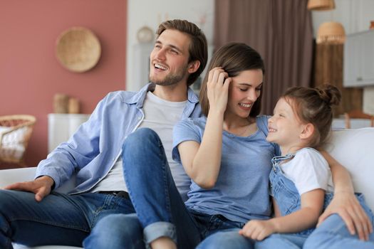 Positive friendly young parents with smiling little daughter sitting on sofa together while relaxing at home on weekend