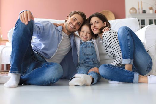 Young Caucasian family with small daughter pose relax on floor in living room, smiling little girl kid hug embrace parents, show love and gratitude, rest at home together