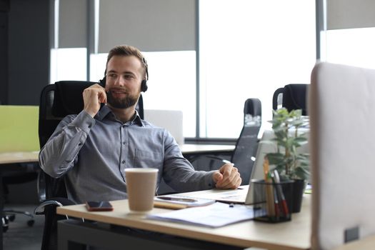 Smiling male call-center operator with headphones sitting at modern office, consulting online information in a laptop, looking up information in a file in order to be of assistance to the client