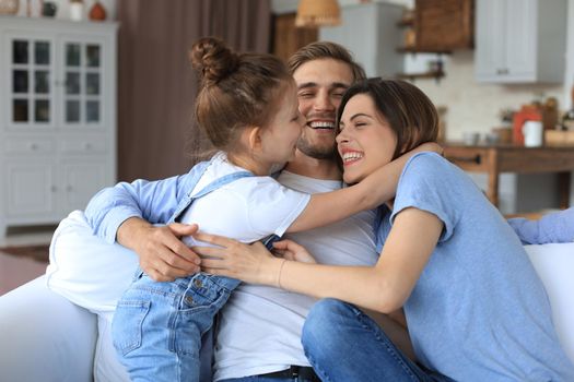 Positive friendly young parents with smiling little daughter sitting on sofa together while relaxing at home on weekend