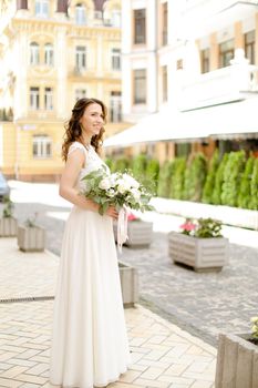 Caucasian happy fiancee walking with bouquet of flowers in city. Concept of bridal photo session.