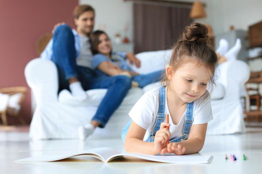 Smiling happy family sit relax on couch in living room watch little daughter drawing in album with colorful pencils. Happy weekends at home