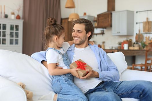 Cute kid daughter make surprise to daddy, little girl presents gift box to father sit on sofa. Father's day