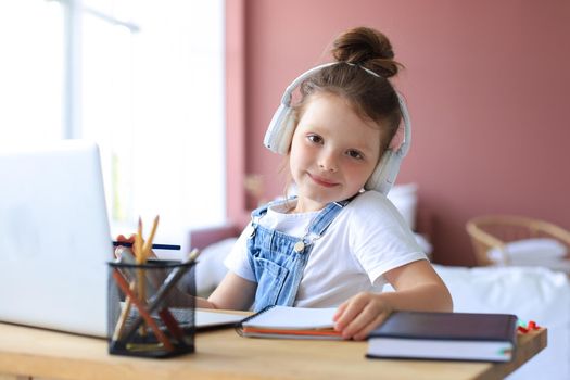 Smiling little girl in headphones handwrite study online using laptop at home, cute happy small child in earphones take Internet web lesson or class on PC