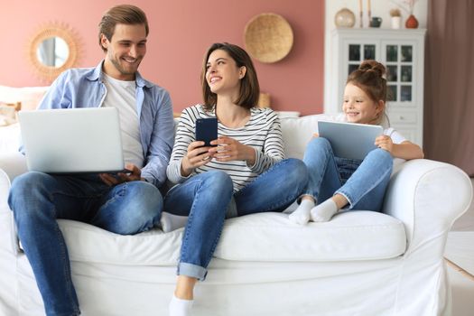 Father, mother and daughter using electronic devices sitting on sofa at living room
