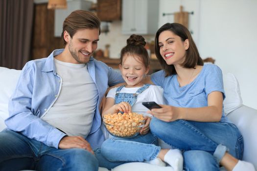 Happy family with child sitting on sofa watching tv and eating popcorn, young parents embracing daughter relaxing on couch together