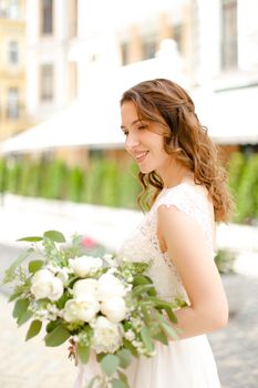 Caucasian smiling fiancee walking with bouquet of flowers in city. Concept of bridal photo session.
