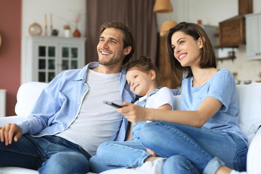 Happy family with child sitting on sofa watching tv, young parents embracing daughter relaxing on couch together