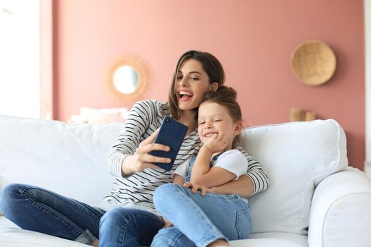 Beautiful young mother and her little daughter taking a selfie with a smartphone at home on the couch
