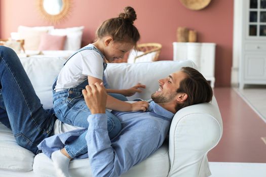 Smiling young father lying on couch at living room and play with happy little daughter