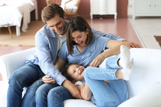 Positive friendly young parents with smiling little daughter sitting on sofa and having fun together at home