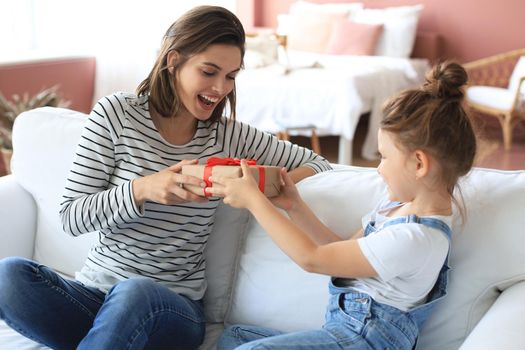 Little girl make birthday present to excited parent while they sitting on couch with mom , smiling little child congratulate give gift box to happy mother