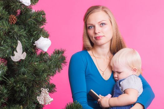 pHolidays, family and christmas concept - young woman with her baby near christmas tree on pink background.