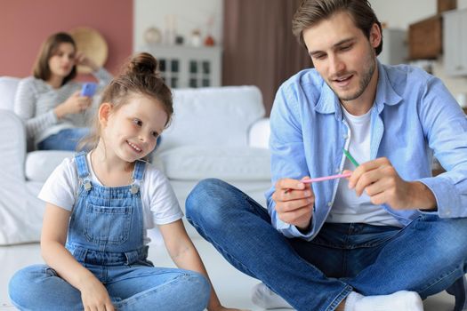 Happy father smilling daughter lying on warm floor enjoying creative activity, drawing pencils coloring pictures in albums, mother resting on couch, family spend free time together