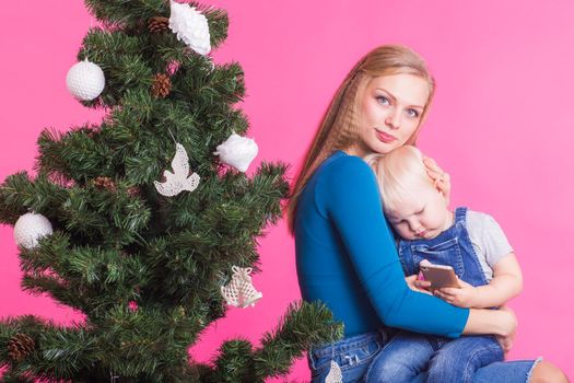 Christmas and holiday concept - Portrait of smiling woman holding her little daughter near Christmas tree on pink background.