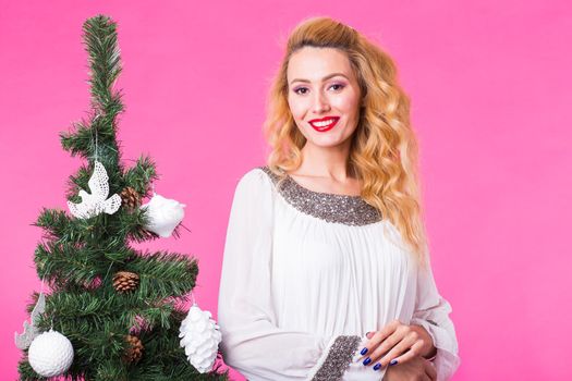 People, holidays and christmas concept - young blonde woman standing near christmas tree on pink background.