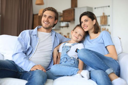 Happy family with child sitting on sofa watching tv, young parents embracing daughter relaxing on couch together
