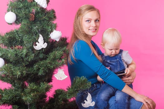 Christmas, holidays, technologies and people concept - young happy woman hugging her daughter near christmas tree. Child using mobile.