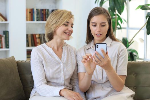 Middle aged mother and adult daughter hugging, using phone together, watching video or photos, sitting on cozy couch at home