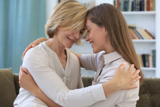 Beautiful middle aged mom and her adult daughter are hugging and smiling while sitting on couch at home
