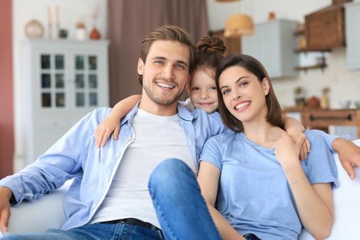 Positive friendly young parents with smiling little daughter sitting on sofa together while relaxing at home on weekend