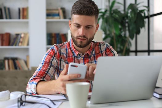 Attractive young man sitting at desk at home office and using mobile phone for cheking social nets