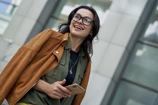Young happy fashionable woman wearing eyeglasses walking on the city street, holding smartphone and looking aside with smile while standing against blurred urban background. People lifestyle concept