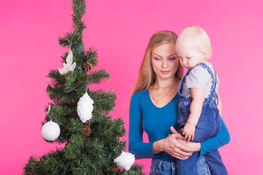 Christmas and holiday concept - Portrait of smiling woman holding her little daughter near Christmas tree on pink background.
