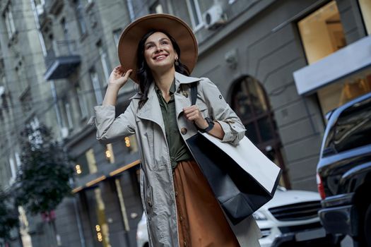 Visiting exclusive boutiques. Young beautiful stylish woman in hat with shopping bag feeling excited, looking aside and smiling while standing on the city street. Fashion, people lifestyle