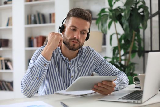 Cheerful young support phone male operator in headset, at workplace while using digital tablet, help service and client consulting call center concept.