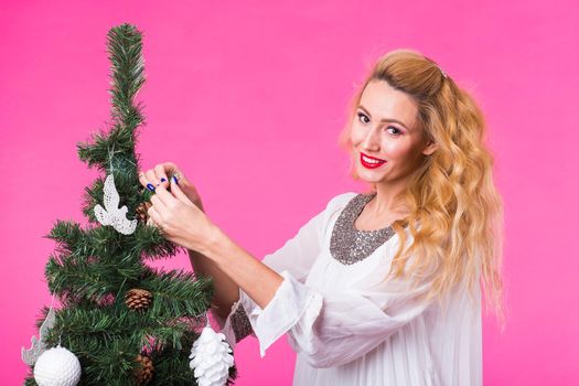 people, holidays and christmas concept - young woman putting christmas ball on christmas tree on pink backgroung.