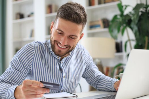 Smiling man sitting in office and pays by credit card with his laptop
