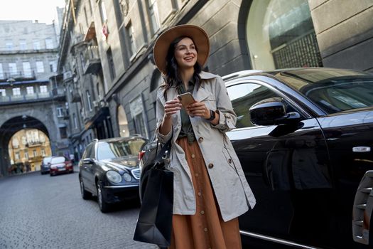 Enjoying shopping. Young happy stylish woman with paper shopping bag using smartphone, looking aside and smiling while standing against black luxury car on the city street. People lifestyle, fashion
