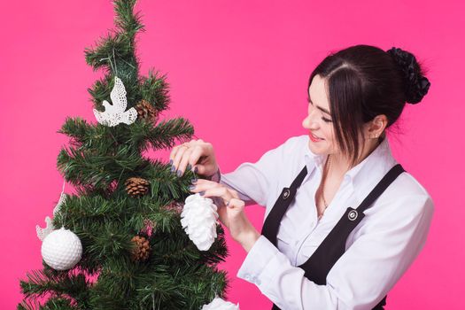 Christmas, holidays and people concept - young happy woman decorating christmas tree on pink background.